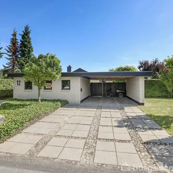 En enkeltfamiliehus med fladt tag står ved en flisebelagt indkørsel. Tallet "12" er synligt på murstensfacaden. Huset er omgivet af velplejede hække og træer.