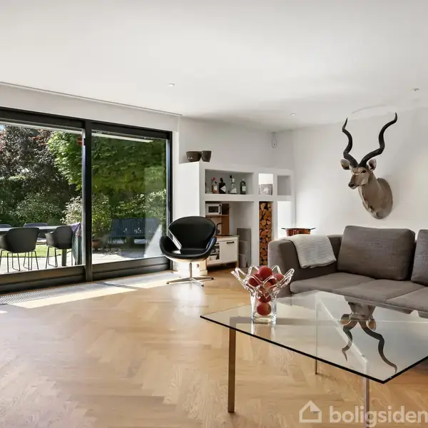 En stue med sofa og glasbord, vægmonteret dyrehoved, store glasdøre, der fører ud til en terrasse med havemøbler, og hylde med dekorationer. Parketgulv og lys indretning.