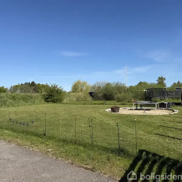 Græsplæne med lille metalhegn i forgrunden. I midten ses en sandfyldt cirkel med en metalstol. I baggrunden er der buske og træer under en blå himmel.