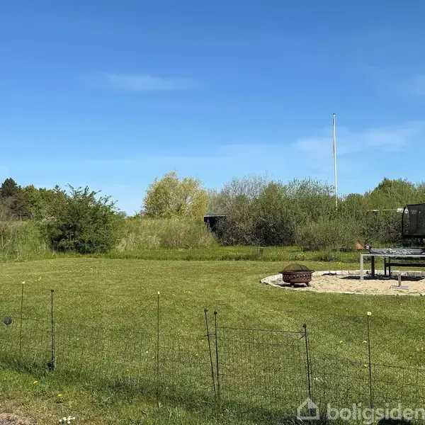 En grøn græsplæne med en brolagt sti ved siden af. I baggrunden ses buske og et hvidt skilt på græsset. Under den blå himmel står en flagstang.
