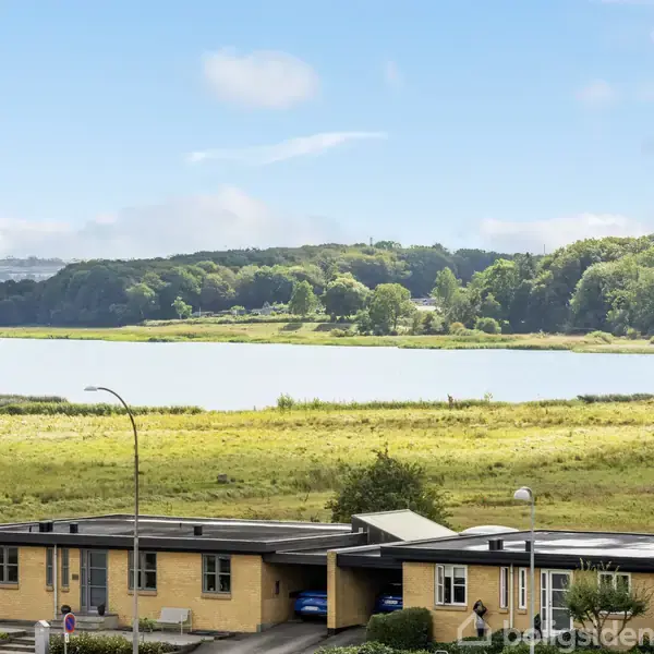 En boligbygning står foran en sø og skov, med blåt himmel i baggrunden. Bygningen er flankeret af gulgrønne marker og en vejlampe.