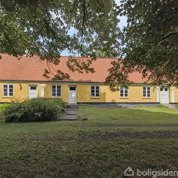 Et gult hus med rødt tegltag står på en græsplæne. Flere døre og vinduer ses foran. Træer omgiver området, hvilket skaber skyggefulde omgivelser.