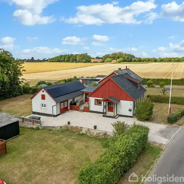 En hvid og rød landejendom med mørkt tag står ved en grusvej. Græsplæne og marker omkranser ejendommen under en blå himmel med skyer.