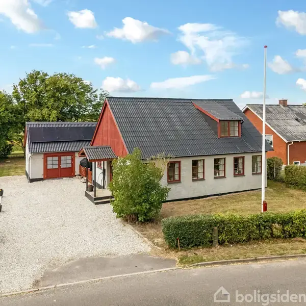 Et hvidt hus med rød gavl og sort tag står stille på en grusindkørsel, omgivet af græs og buske, ved en vej.