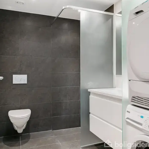 Toilet hænger på mørk flisevæg, tøjvaskemaskine-tørretumbler kombination står til højre, og hvide skuffer ses under en vask i et moderne badeværelse.