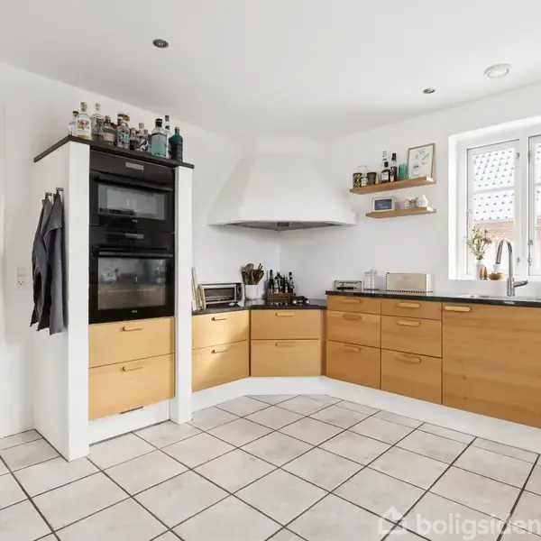 Et køkken med lyse træskabe og hvide vægge. Skabet har indbyggede ovne, og der står flasker ovenpå. Kokkenet har hvide fliser, vindue med planter, og redskaber på bordpladen.