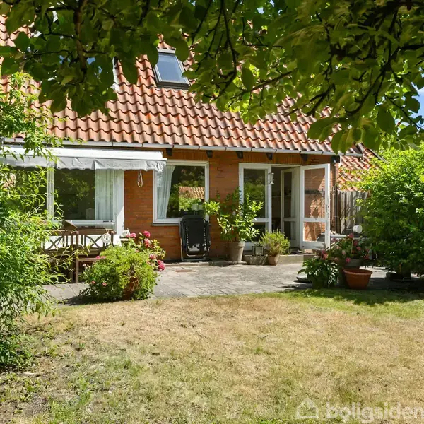 Hus med rødt tegltag og murstensvægge, omgivet af en have med en terrasse. Haven har grønne buske, træer og en græsplæne. En bænk og nogle krukker med planter står på terrassen.