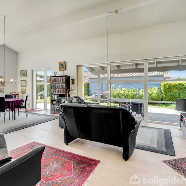 En stue indrettet med sorte lædersofaer står vendt mod et glasudsnit, der tilbyder udsigt til en have. En spiseplads med lilla stole ses til venstre.