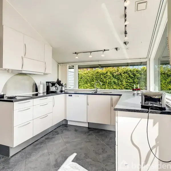 Et moderne køkken med hvide skabsfronter og mørke bordplader. Det er veludstyret med apparater som en kaffemaskine og brødrister. Store vinduer giver udsigt til en grøn hæk.