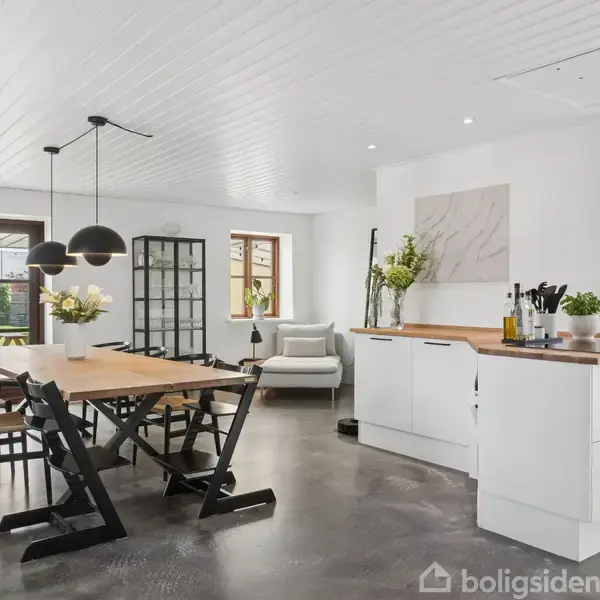 Køkken med hvidt interiør og trægulv; spisebord med sorte stole; køkkenø med planter og redskaber; store vinduer og dør til have. Ingen tekst findes udover "Boligsiden".