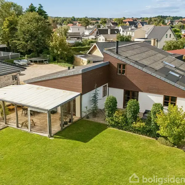 En moderne bungalow med skråt tag, store vinduer og en overdækket terrasse i en frodig have. Omkranset af flere huse i et roligt nabolag med træer og grønne områder.