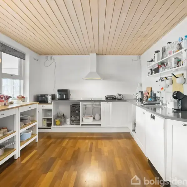 Køkken med hvide skabe og træbordplader, kaffemaskine og brødrister, åben hylde med krydderier, træloft og vindue med moderne lampe. Kaffe og kiks på bordet til venstre.