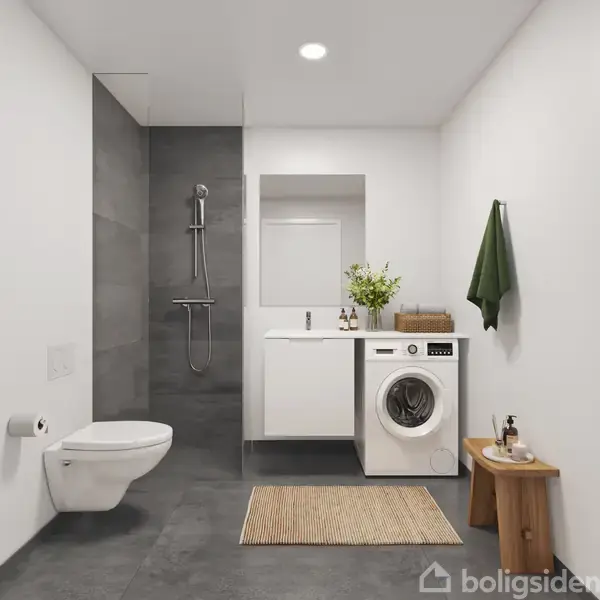 Toilet hænger på væggen; ved siden af er en bruser. Vaskemaskine under vask, med spejl over, står i et lyst badeværelse. Grøn håndklæde hænger på krog.