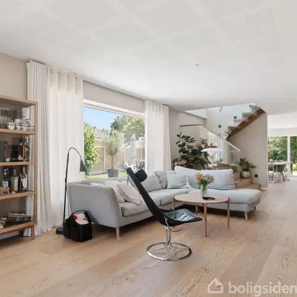 En lys stue med en grå sofa og sort stol står ved et stort vindue. Hyggelige planter og hylder spækket med genstande omkranser rummet, mens en trappe fører opad.