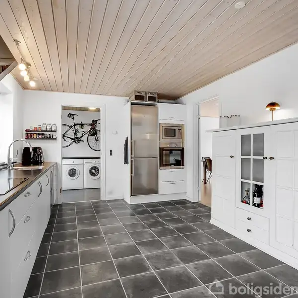 Et lyst køkken med hvide skabe og gråt flisegulv. I baggrunden ses en vaskemaskine og tørretumbler ved siden af en cykel monteret på væggen.