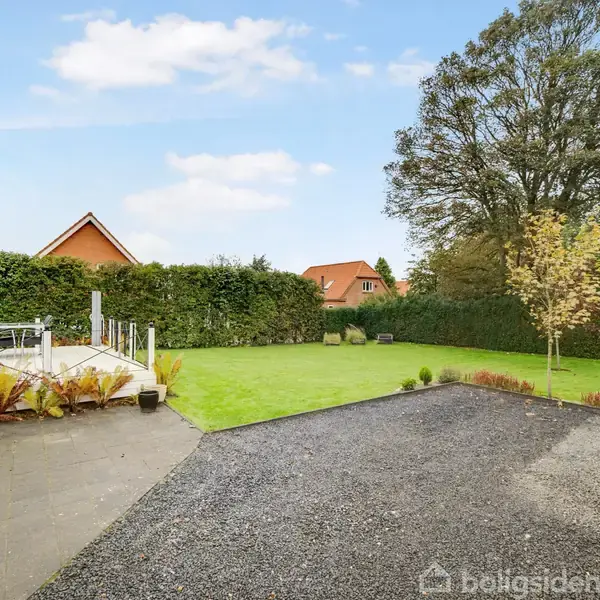 En grusbelagt indkørsel fører til en rummelig græsplæne med træer i en villaområde. En terrasse med havemøbler er til højre. Baggrunden viser klare somrelementer.
