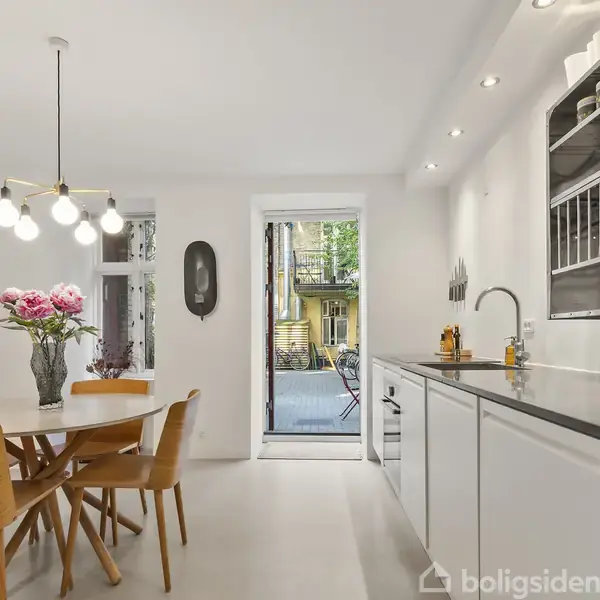 Et køkken med moderne indretning, hvor et rundt spisebord med blomster står i midten. Til højre er der køkkenbord og overskabe. Bagved er der en dør til en terrasse.