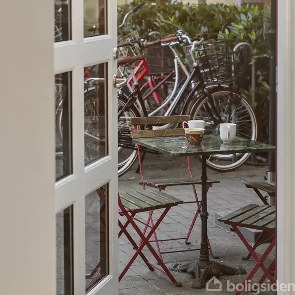 En cafébord med to krus står udenfor en glasdør. Omkring bordet er der to stole, og cykler er opstillet i baggrunden.