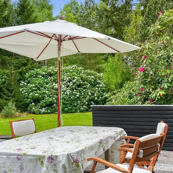 En terrasse med et bord dækket af en mønstret dug og fire stole. En stor parasol står midt på bordet. Omgivet af grønne planter og blomstrende buske.