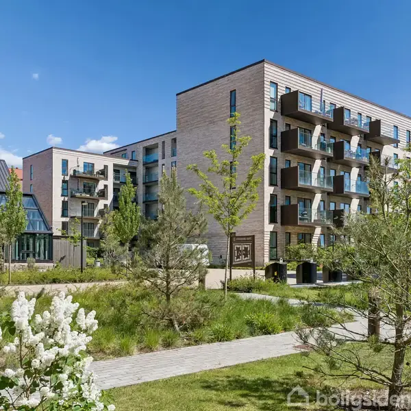 Flere moderne lejlighedsbygninger med altaner står i en grøn have. Blå himmel og enkelte skyer i baggrunden danner rammen omkring de frodige omgivelser.