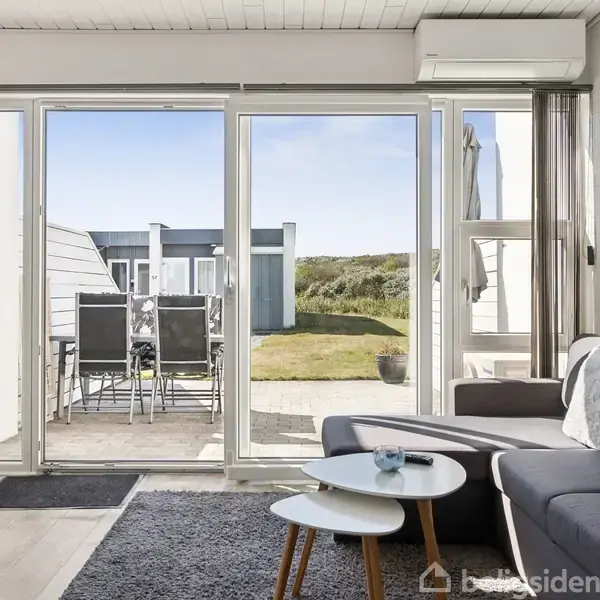 Stue med grå sofa og sofabord i forgrunden. Store glasdøre fører ud til terrasse med bord og stole. Grøn græsplæne og moderne bygninger ses i baggrunden.