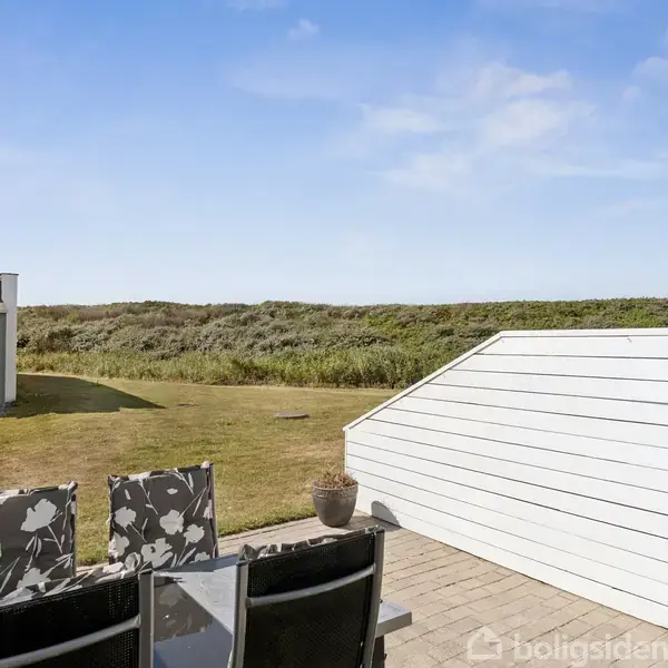 Terrassemøbler står stille ved et bord på en flisebelagt terrasse. I baggrunden ses et grønt græsområde og en lav bevoksning. Himlen er klar og solrig.