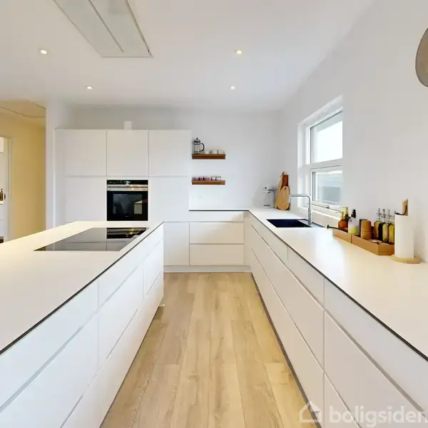 Et moderne køkken med hvide skabe og en indbygget ovn. Køkkenøen har en kogeplade. Rummet har trægulv, stort vindue og en vægmonteret ur.ippetspolis