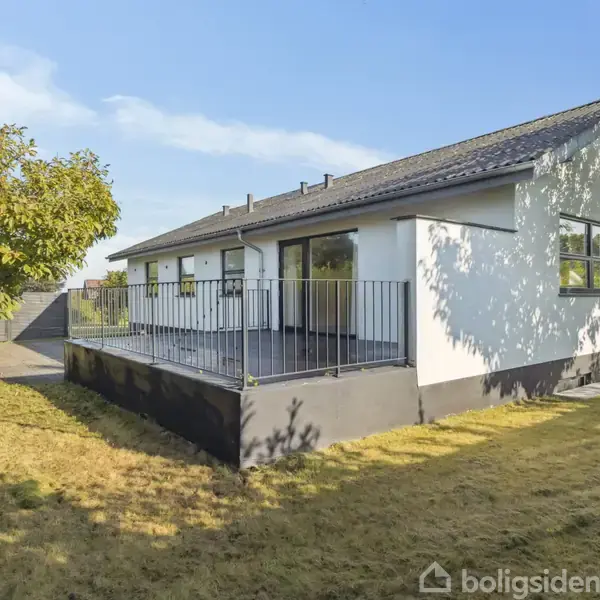 En hvid en-etagers hus står på en græsplæne med et træ ved siden af. Huset har en terrasse med sort gelænder. I baggrunden ses flere træer og en klar himmel.