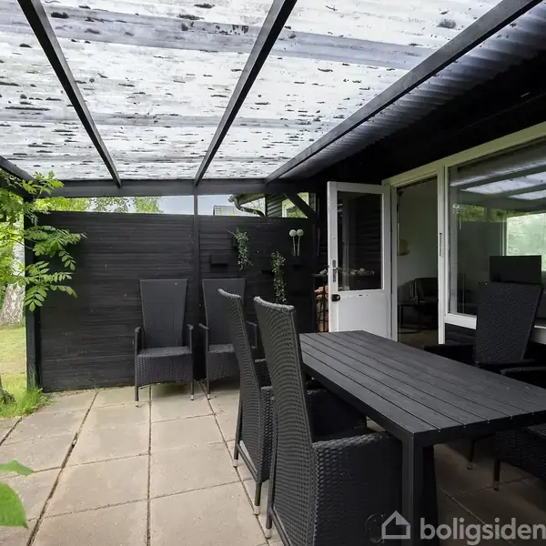 Terrasse med sort bord og stole i flet. Handlingen sker under et gennemsigtigt tag. Haven er synlig til venstre med træer og græsplæne i baggrunden.