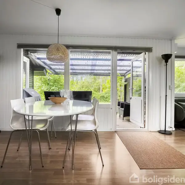 Et rundt spisebord med stole står i en lys stue. Store vinduer og en åben dør viser en terrasse med grønne omgivelser. En sofa er placeret til højre.