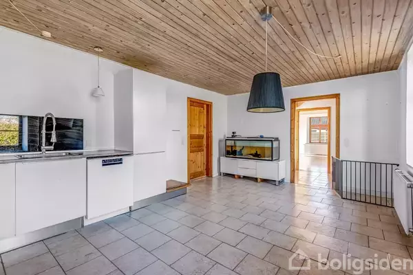 Et moderne køkken med hvide skabe står stille. Stue med lyst trægulv og loft; sort pendellampe hænger. Et akvarium står på et skab ved trædør.