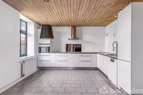 Køkken med hvide skabe og indbygget ovn har et trægulv og -loft. En sort lampe hænger fra loftet, og et stort vindue giver naturligt lysindfald.