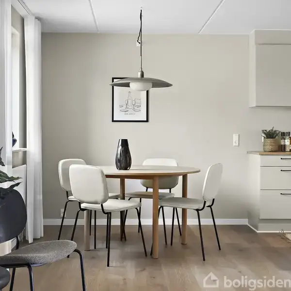Rundt spisebord med hvide stole er centralt placeret, omgivet af minimalistiske møbler og beige vægge. En sort vase står på bordet, og en pendellampe hænger over det.