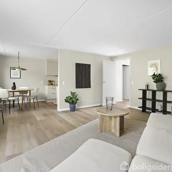 En lys stue med en beige sofa i forgrunden. Et rundt træbord står foran sofaen. Bagved ses et spisebord og stole i et åbent køkkenområde. Planter pynter rummet.