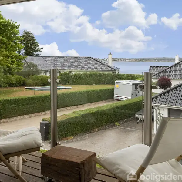 To terrassestole står på en altan med udsigt over en have og hustage. En hæk adskiller terrassen fra en indkørsel. I baggrunden ses en sø og skyer.