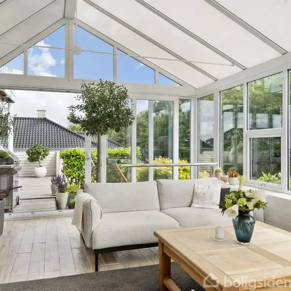 To udendørs stole står på en grusbelagt terrasse ved siden af et moderne drivhus med glasvægge og loungemøbler. Omgivet af grønt løv og en velplejet græsplæne.
