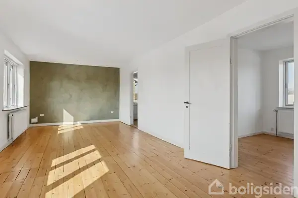 En lys stue med trægulve og hvide vægge har en grøn accentvæg. Døren står åben til et tilstødende rum. Vinduer lader sollys skinne ind, skaber skygger på gulvet.