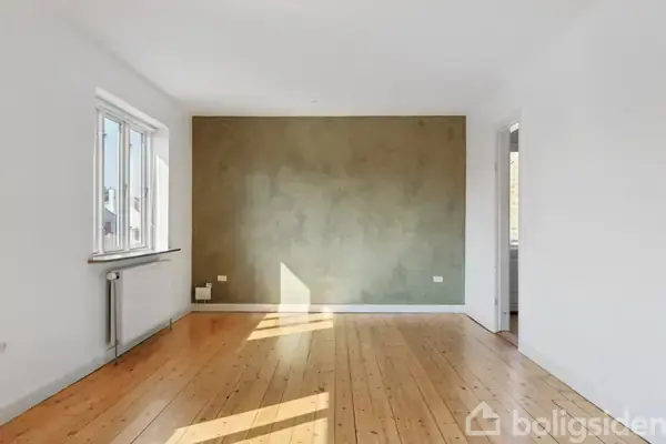 Et lyst rum med trægulv og en olivengrøn væg. Sollys strømmer ind fra et vindue til venstre, og der er en radiator under vinduet.