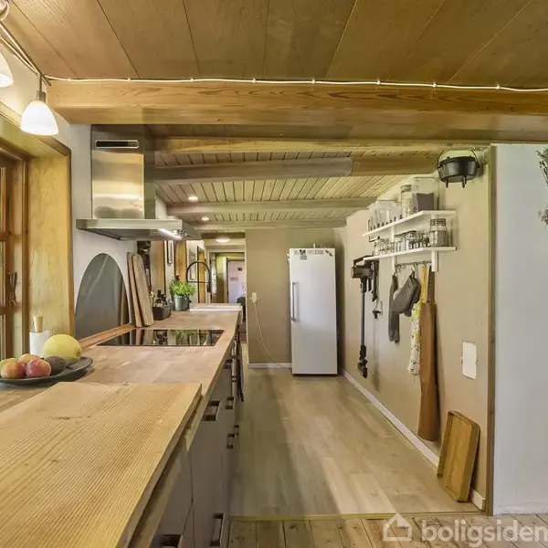 Et rummeligt køkken med trægulv og -loft har en køkkenø med frugter, en lang bordplade, træhylder med køkkenudstyr, og et hvidt køleskab. Store vinduer giver udsigt til det grønne udenfor.