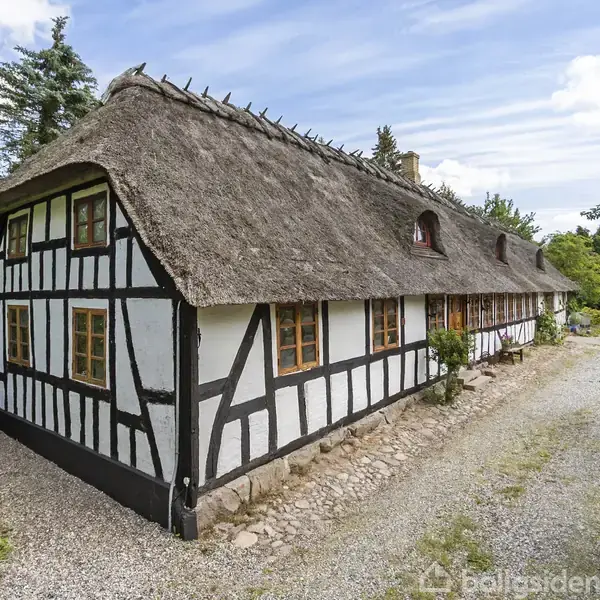 Bindingsværkshus med stråtag står langs en grusvej; huset har hvide og sorte vægge med små vinduer. Omgivet af grønt løv, under en blå himmel med skyer.
