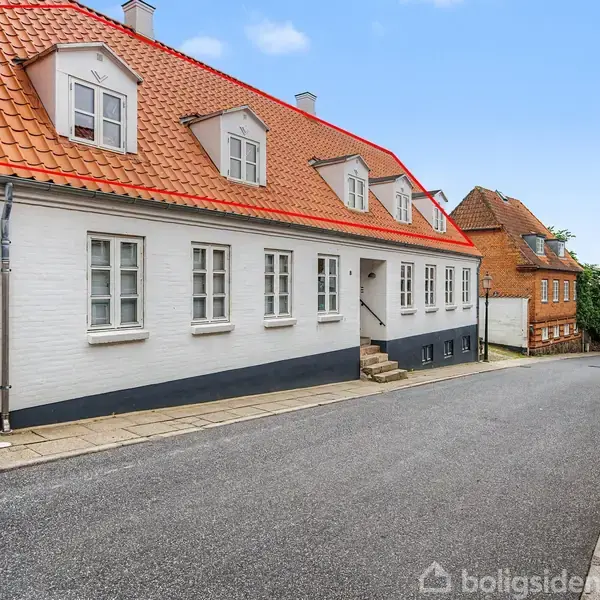 Hvid murstensbygning med rødt tegltag og små kviste, står på en skrånende vej med andre murstensbygninger, himlen er blå.