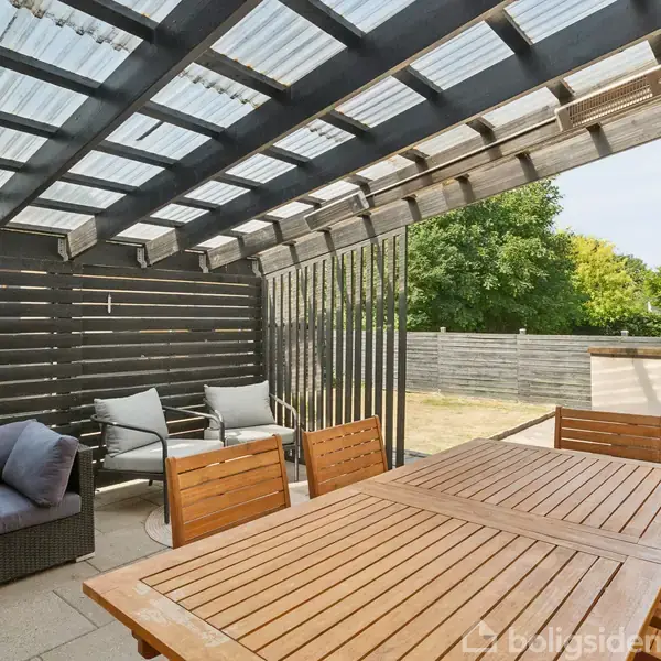 En overdækket terrasse med et stort træbord omgivet af stole. I hjørnet står en hjørnesofa med grå puder. Hegnet omkranser terrassen, og der er grønne træer i baggrunden.