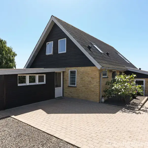 Et trekantet hus med sorte tagsten står på en brostensbelagt gårdsplads. Murstensvæggene er gule, og der er vinduer med hvide rammer. En busk vokser tæt på huset.