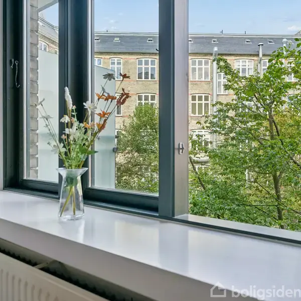 En glasvase med blomster står på en vindueskarm, mens den udsigt mod en bygning og træer udenfor.