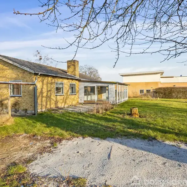 Et gammelt murstenshus står ubenyttet. Græsset vokser vildt omkring, mens en solbadet have omkranser bygningen. En afskåret træstub står nær en flisebelagt sti under blå himmel.