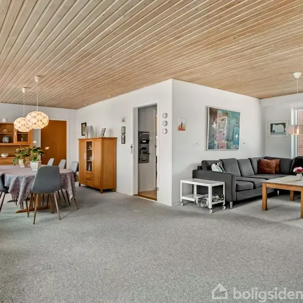 Sofa og spisebord står i en lys stue med træloft og gråt tæppe. Rummet har vinduer, malerier, reoler og hængende lamper, der skaber en hyggelig atmosfære.