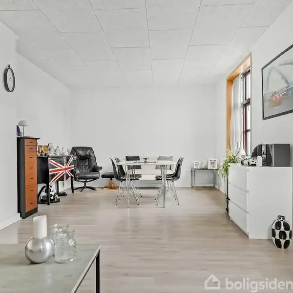 Stue med et spisebord og stole, en kontorstol ved et skrivebord, og en hvid kommode. En væg klokke og vægbilleder dekorerer det lyse rum.