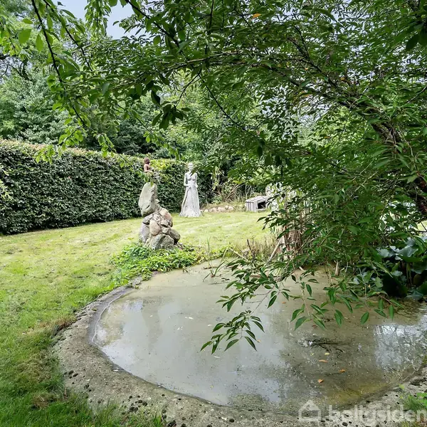 En lille dam med klart vand er omgivet af grønne træer og buske i en velplejet have. To havefigurer står nær dammen, som skaber en fredelig atmosfære.