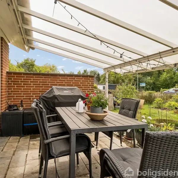 En overdækket terrasse har et spisebord med seks stole og en grill. Omkring terrassen er der en have med græsplæne og buske.