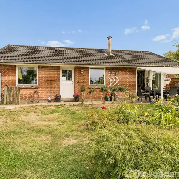 Et rødt murstenshus med sort tag står omgivet af en have. Der er en terrasse med møbler til højre og blomster foran huset. Himlen er blå og klar.